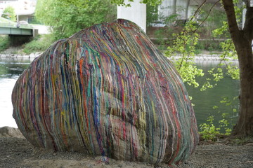 a boulder on a river bank painted with thin striped of different colors with. A tree trunk on the...