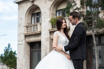 Fototapeta premium Bride and groom hugging in the old town street. Wedding couple walks in Budapest near Parliament House. Caucasian happy romantic young couple celebrating their marriage. Wedding and love concept.