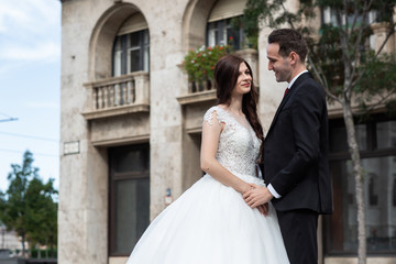 Fototapeta premium Bride and groom hugging in the old town street. Wedding couple walks in Budapest near Parliament House. Caucasian happy romantic young couple celebrating their marriage. Wedding and love concept.