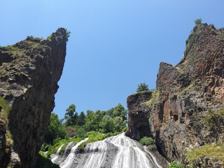 From a height, water flows down. Beautiful waterfall between the rocks. Behind the waterfall there are trees and behind them the blue sky.