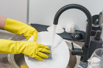 Close up on yellow rubber protective gloves on female unknown woman woman's hands manually washing plates dishes in the kitchen by the sink side view at home by the tap water in day side