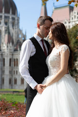 Naklejka premium Bride and groom hugging in the old town street. Wedding couple walks in Budapest near Parliament House. Caucasian happy romantic young couple celebrating their marriage. Wedding and love concept.