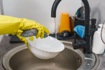 Close up on yellow rubber protective gloves on female unknown woman woman's hands manually washing plates dishes in the kitchen by the sink side view at home by the tap water in day side