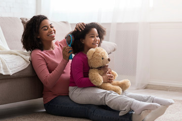Caring pregnant african mom combing her little daughter's hair at home