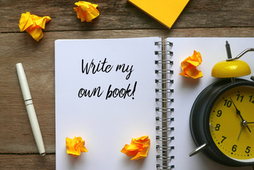 Top view of table clock,trash paper,sticky notes,pen and notebook written with ' Write my own book! ' on wooden background.