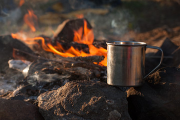 Metal camping mug with hot tea by the fire in the forest. Trekking, camping dishes, bonfire, evening