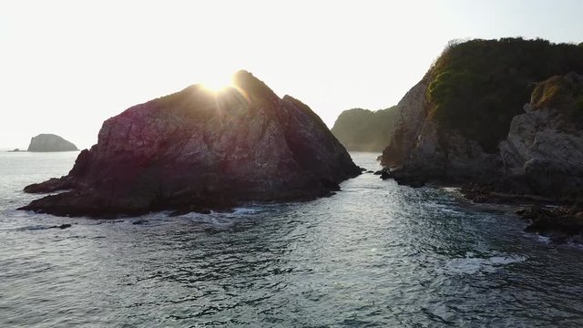 Drone flies low and close between two rocky coastal cliffs towards the setting sun.