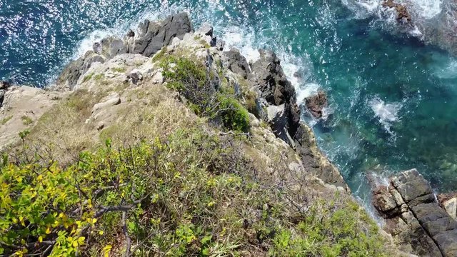 Drone flies at close proximity off a cliff in the tropics looking straight down towards the crashing surf and then flies towards the Ocean with the camera looking up towards the horizon.
