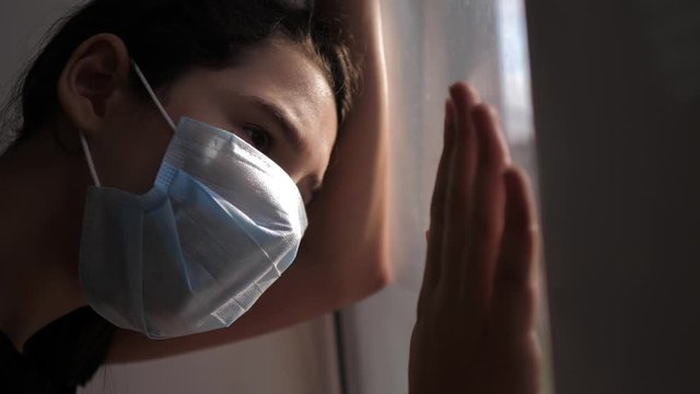 little girl looks out the window sad in a medical lifestyle gauze mask. concept pandemic virus covid 19 self-isolation infection doomsday. the girl touches the glass sunlight wants to go outdoors