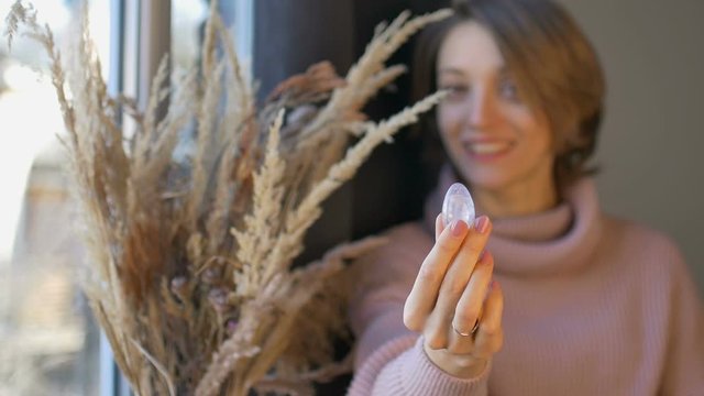 Woman is holding yoni egg made from transparent violet amethyst stone standing near the window with vase of spikelets. Female health concept, vumfit, imbuilding or meditation
