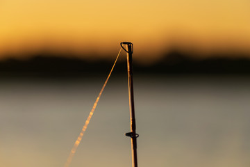 Closeup of fishing rod tip silhouetted against evening sunset and water with plenty of copy space.