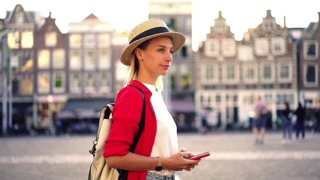 Slow motion effect of Caucasian young female standing outdoors on square with beautiful architecture of historic town, Hipster girl tourist using smartphone while visiting in city during vacations
