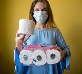 Bialystok / Poland-30.04.2020: girl in protective medical mask and disposable gloves stands with a toilet paper in her hands on a plain yellow background. Panic and self-isolation due to coronavirus