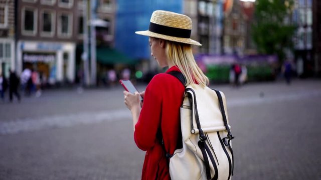 Slow motion of millennial female tourist with backpack messaging with people on travel blog using 4g internet connection outdoors in city.Hipster girl taking photo for share media in social network

