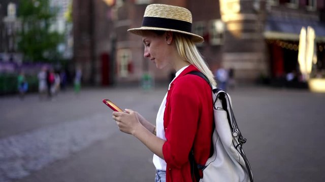 Happy caucasian smiling hipster girl with backpack typing message in application using cellular camera for taking images of city sightseeing during travel. Millennial female tourist enjoying trip
