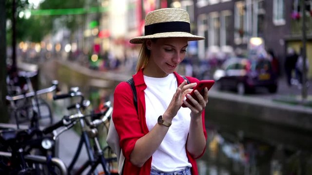 Slow motion of caucasian female tourist spending vacations in Amsterdam city searching new sightseeing on browsing websites via cellphone, young traveler standing with mobile phone in hands
