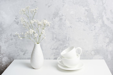 Bouquet of small delicate flowers gypsophila in vase and and two white cups for tea or coffee on table agains gray concrete wall. Copy space Front view