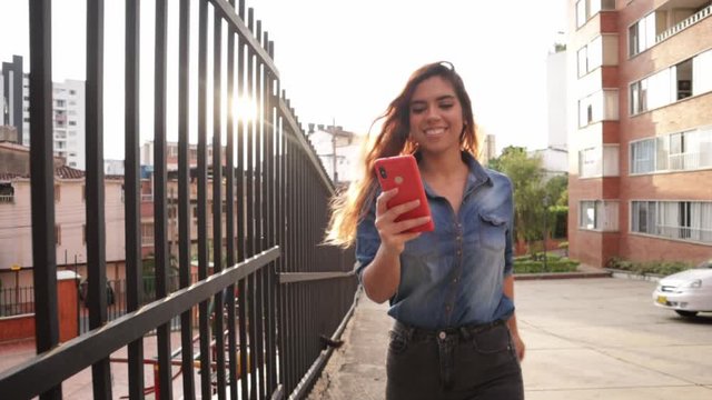 Happy latin woman walking with cell phone in hand and sunset