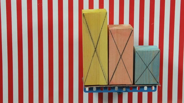 Colorful wood blocks of different size placed on a board that gradually tilts, one by one they fall as their marked centrum of gravity exceeds their base. The background lines are vertical reference, 