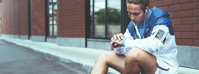 Young athlete in blue windrunner sitting on the street adjusting running program for his morning workout using smart watches on his hand. Wide screen, panoramic