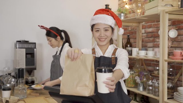 smiling young female barista in apron holding coffee to go in cup and take away food in paper bag face camera. two cafe bar staff girls wearing santa hat in christmas holidays working in store.