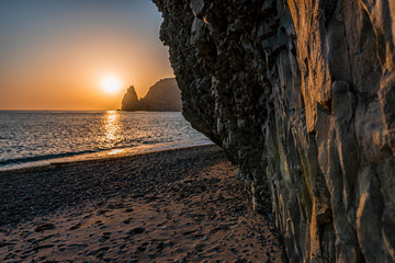 view of the sunset sea and the beach, the volcanic rock is lit by the warm sunset, sand and...
