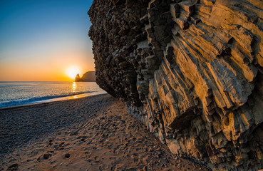 view of the sunset sea and the beach, the volcanic rock is lit by the warm sunset, sand and...
