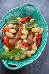 Green serving tray with baked potato gnocchi, pork sausages and bell peppers, vertical shot on a grey concrete background