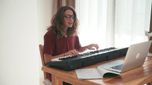 A young woman singing and playing the piano keyboard online using her laptop. Concert for friends in a COVID-19 self-isolation time.