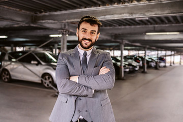 Young successful bearded auto dealer in suit standing on parking lot with arms crossed and looking at camera.