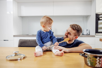 Fototapeta premium Adorable blond little boy sitting on kitchen counter and holding mixing spoon. Father crouching next to him. They are preparing meal for family.