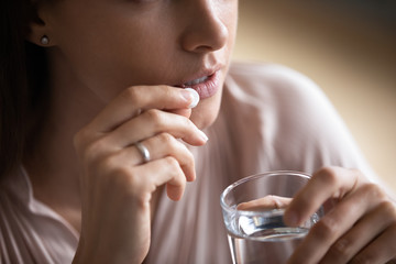 Close up young woman taking white round pill, holding water glass, unhealthy female taking...