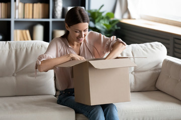 Smiling young woman unpacking parcel, sitting on couch at home, happy attractive female looking in...