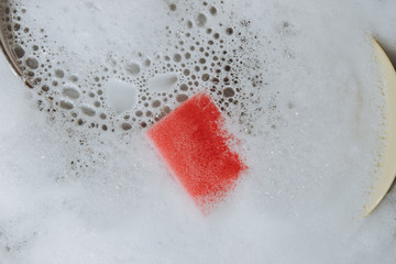 Красная губка лежит в раковине полной пены . Уборка кухни. Мыльная вода для мытья посуды . 