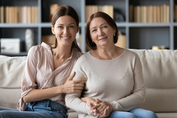Portrait of smiling adult grown up daughter and middle aged mother sitting on cozy sofa at home, posing for family photo together, looking at camera, young woman with mature mum holding hands