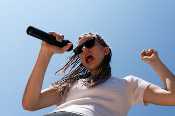 White teenager with hair braids wearing a white top and sun glasses singing a song with a black microphone at the right hand and the sky and the sunlight at the background. Horizontal photo