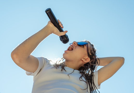 White teenager with hair braids wearing a white top and sun glasses singing a song with a black microphone at the right hand and the sky and the sunlight at the background. Horizontal photo
