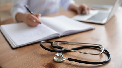 Female doctor physician sits at workplace using laptop, writing notes in medical journal. Woman...