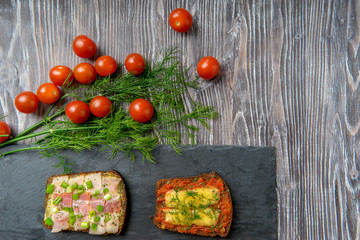 Sandwiches made from slices of rye bread with bacon and cheese on dark stone and wooden surface are served with fresh green dill and red cherry tomatoes top view. Background with delicious snacks