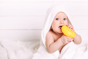 A cute baby with blue eyes wrapped in a white towel as if in a hood holds a yellow rubber toy duck. Clean baby after bath, taking care of children. Copy space