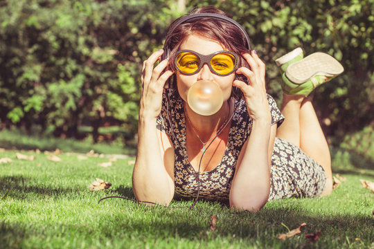 Happy girl with headphones listen to music and  inflates a chewing gum. Stylish young woman in casual style and  yellow sunglasses outdoors 