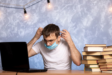 young man takes off a medical mask while sitting at a table with books and a laptop. quarantine end. infection protection. work in the office.