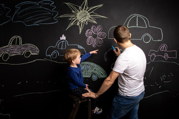 a little boy stands on the stairs near the slate wall and draws cars with his dad with chalk....