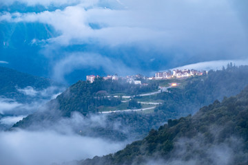 Fototapeta na wymiar Hotel in the mountains among the green forest. Beautiful clouds and heavy fog at sunset in the mountains. Krasnaya Polyana, Sochi, Russia