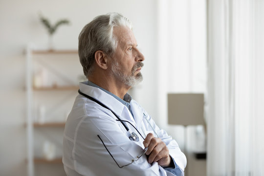 Thoughtful serious senior doctor looking through window lost in thoughts. Worried pensive old physician thinking of healthcare question, concerned of challenge, feels anxious makes difficult decision.