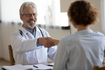 Friendly smiling professional physician handshaking female patient at medical consultation. Happy...