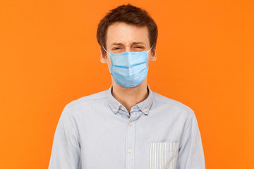 Portrait of sad alone young worker man with surgical medical mask standing and looking at camera with upset frown face. indoor studio shot isolated on orange background.
