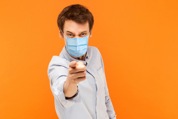 Hey you. Portrait of angry young worker man with surgical medical mask standing pointing and scolding at camera with mad face. indoor studio shot isolated on orange background.