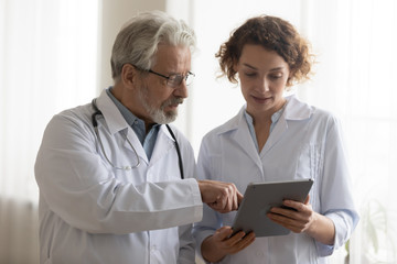 Medical team of two professional doctors talking and using digital tablet. Senior male chief physician helping young female nurse holding medical tech device discussing patient diagnosis in hospital.