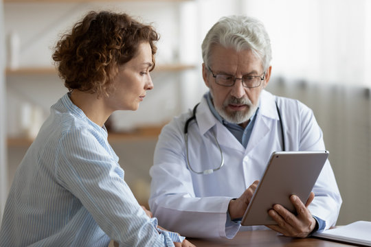 Older adult doctor consulting young female patient using digital tablet tech during checkup appointment meeting. Woman client visiting physician listening diagnosis treatment prescription in hospital.